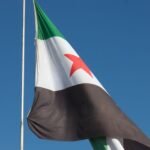 Syrian flag with red star waving on a pole against a clear blue sky.