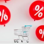 Mini shopping cart with red percentage balloons on a white background, symbolizing sales.